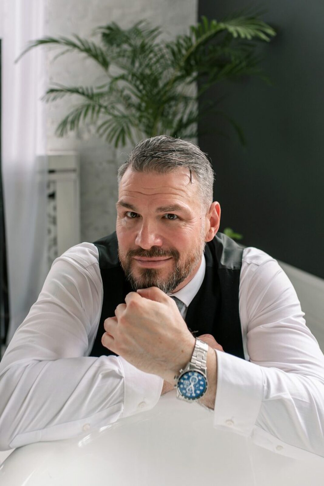 A young man with dark hair and blue eyes, wearing a light blue dress shirt and black pants, sitting on a chair in an indoor setting with large green plants around him and a window in the background.
