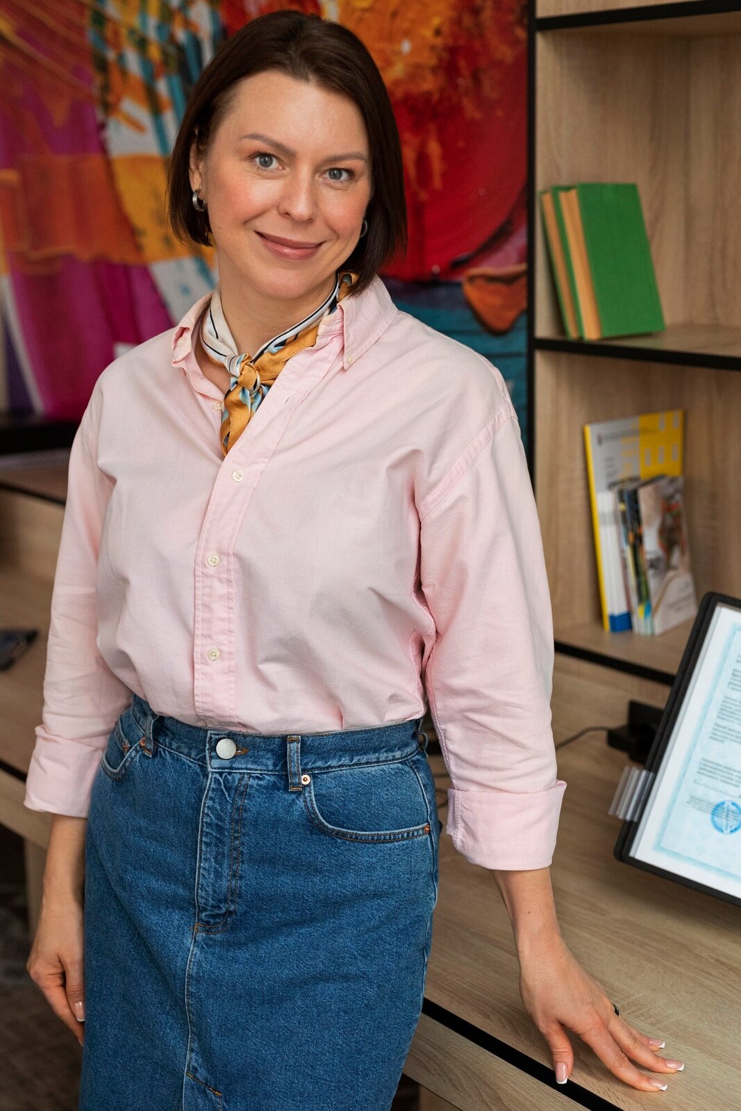 A young woman with long brown hair wearing a black dress with puffed sleeves, standing indoors near large green plants, with a blurred background showing a window and a door.